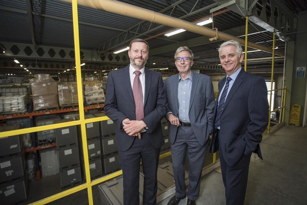 3 men in suits smiling in a warehouse with stacks of boxes in the background