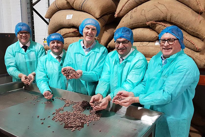 5 men wearing hair nets and overalls while holding cocoa beans
