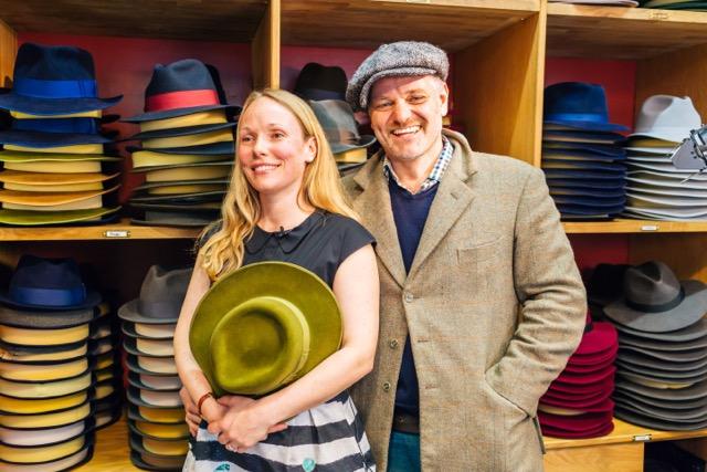 A man and a woman in a hat shop holding a hat with stacks of hats in the background