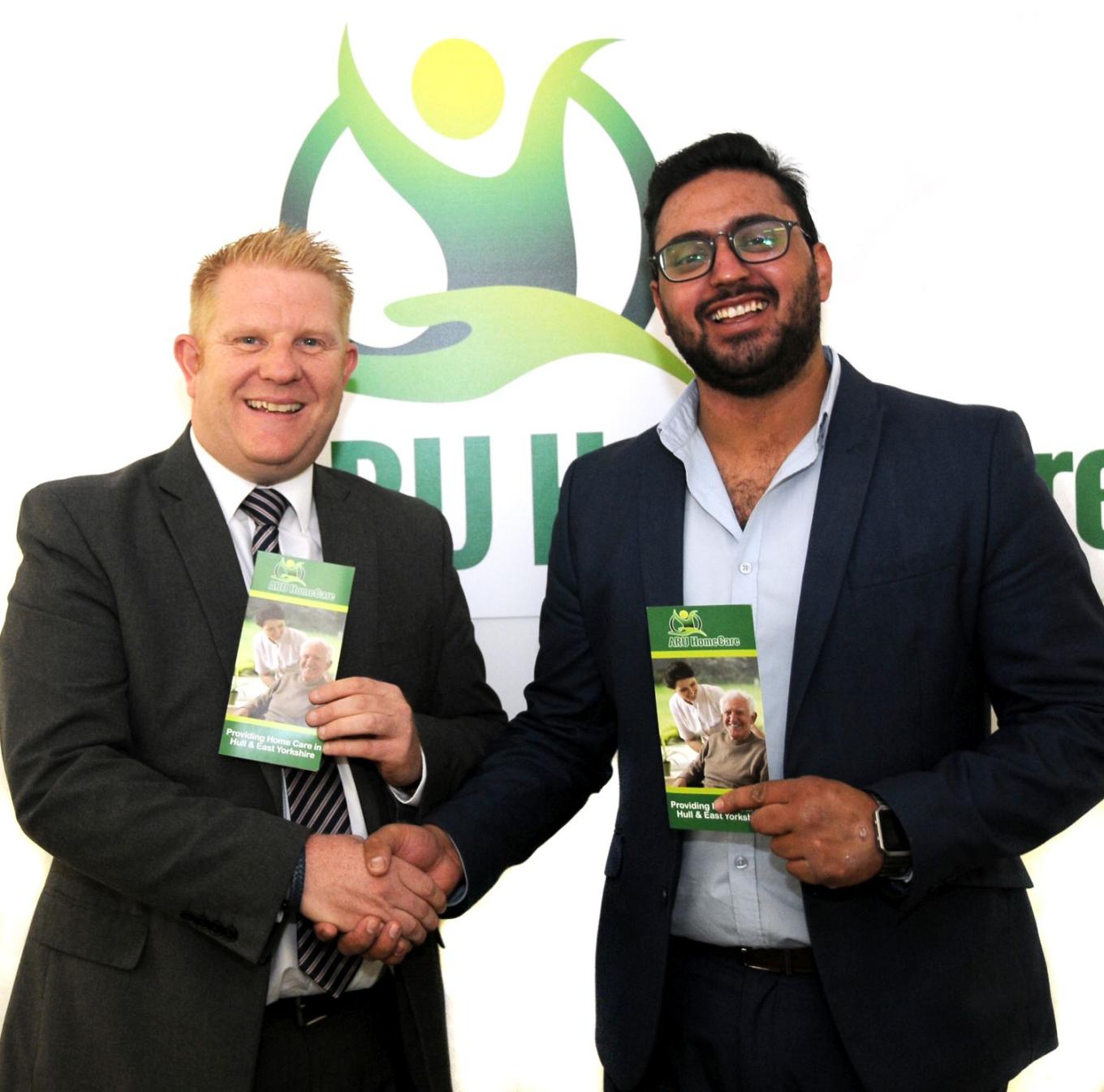 2 men in suits shaking hands and holding Aru Home Care leaflets with the logo on the wall behind them