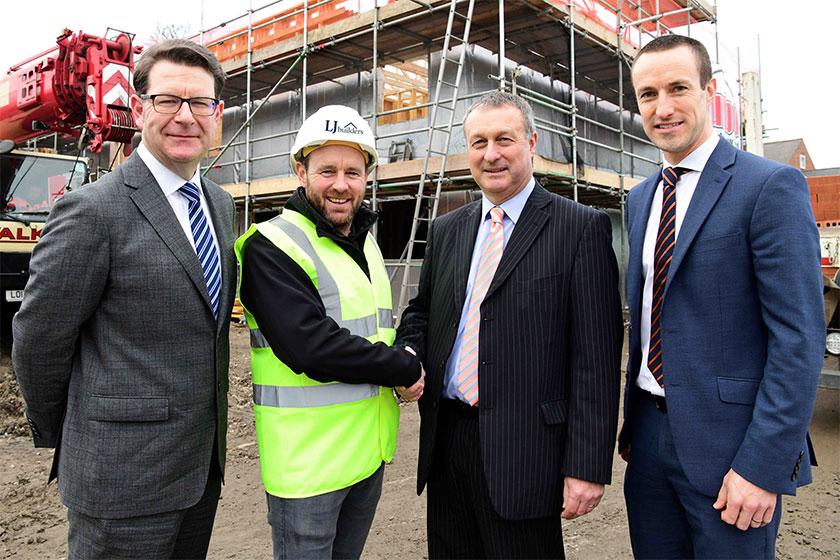 3 men in suits and one man in a Hi - Vis jacket stood in front of a construction site