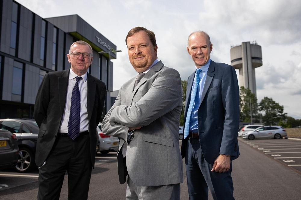 3 men in suits smiling and stood in a car park outside an office building