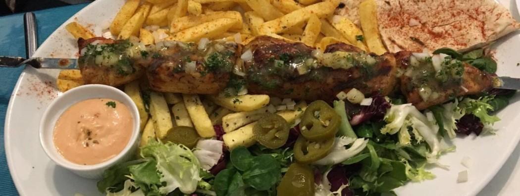 A close up of a Greek chicken skewer, chips, salad and sauce