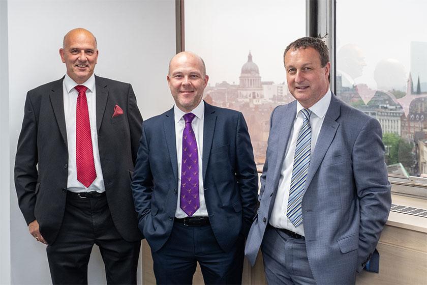3 men in suits smiling and stood in a meeting room with the city of London in the background