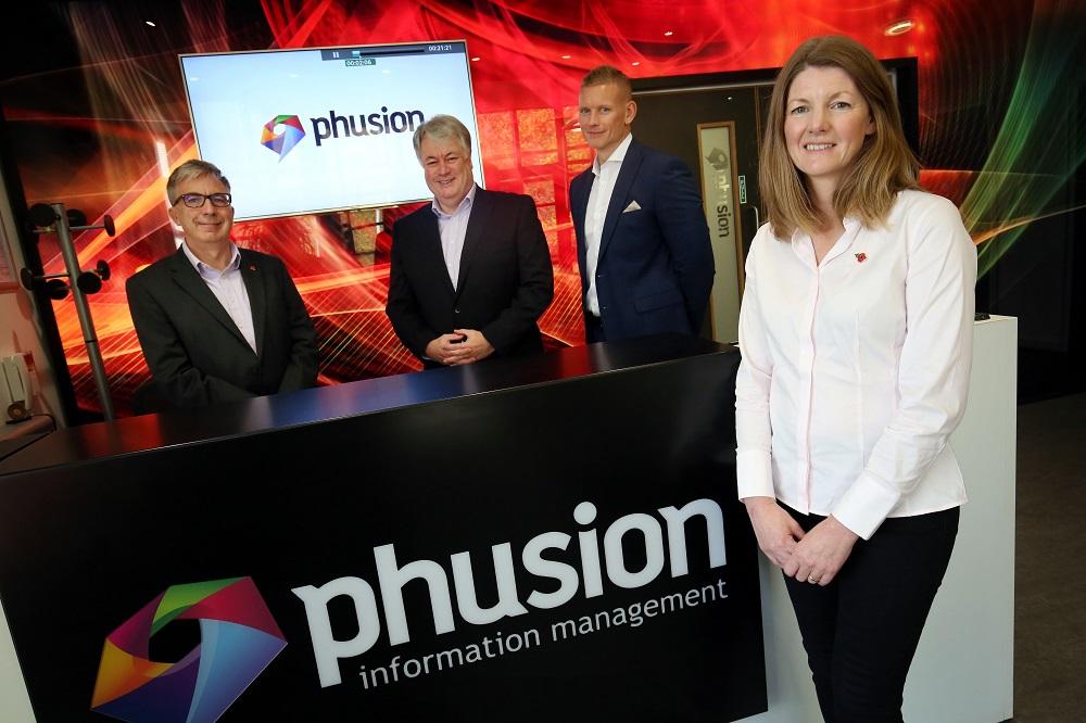 3 men and a woman stood near a Phusion branded counter and TV screen