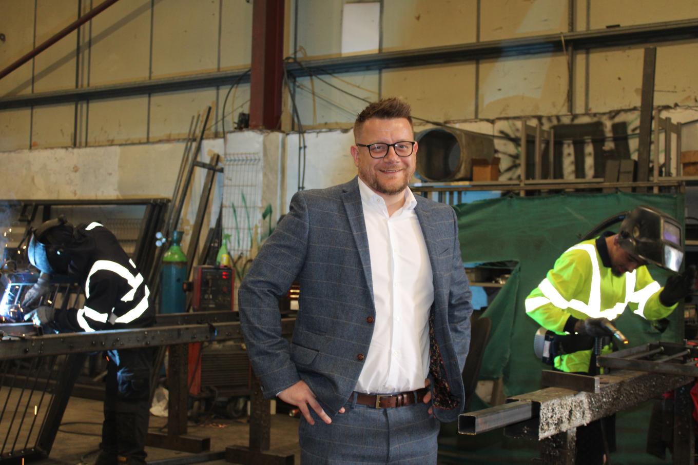 A man wearing a suit stands before a metal shop, embodying a professional presence in an industrial environment